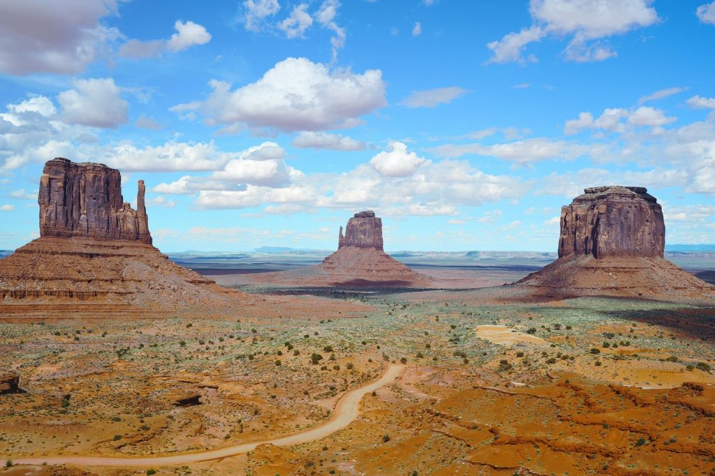 A winding desert road leads through Monument Valley’s iconic red buttes under a bright blue sky filled with scattered clouds.