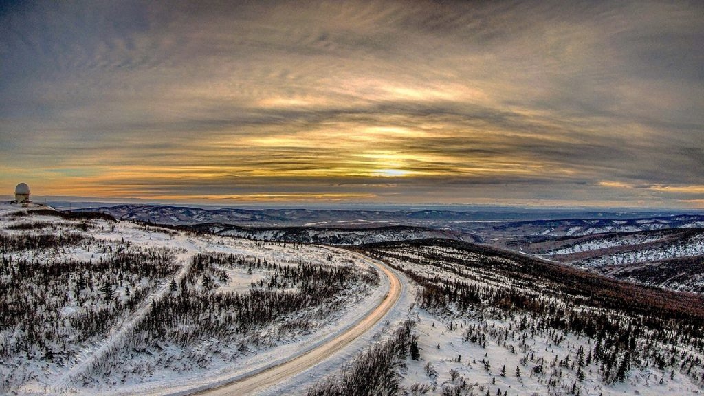A snowy road winds through Alaskan hills under a glowing sunset sky.