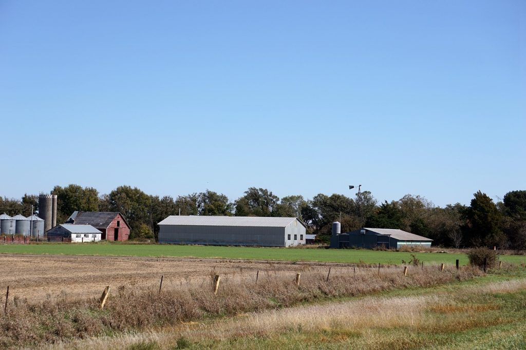 A Kansas farm stretches across flat fields under a clear blue sky, with barns, silos, and trees dotting the rural landscape.