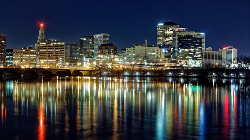 Hartford’s city skyline glows at night, with colorful lights reflecting off the Connecticut River and a bridge adding to the vibrant, peaceful scene.