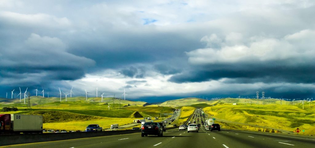 A busy highway winds through green hills under dramatic clouds, with wind turbines lining the landscape, blending travel with renewable energy.