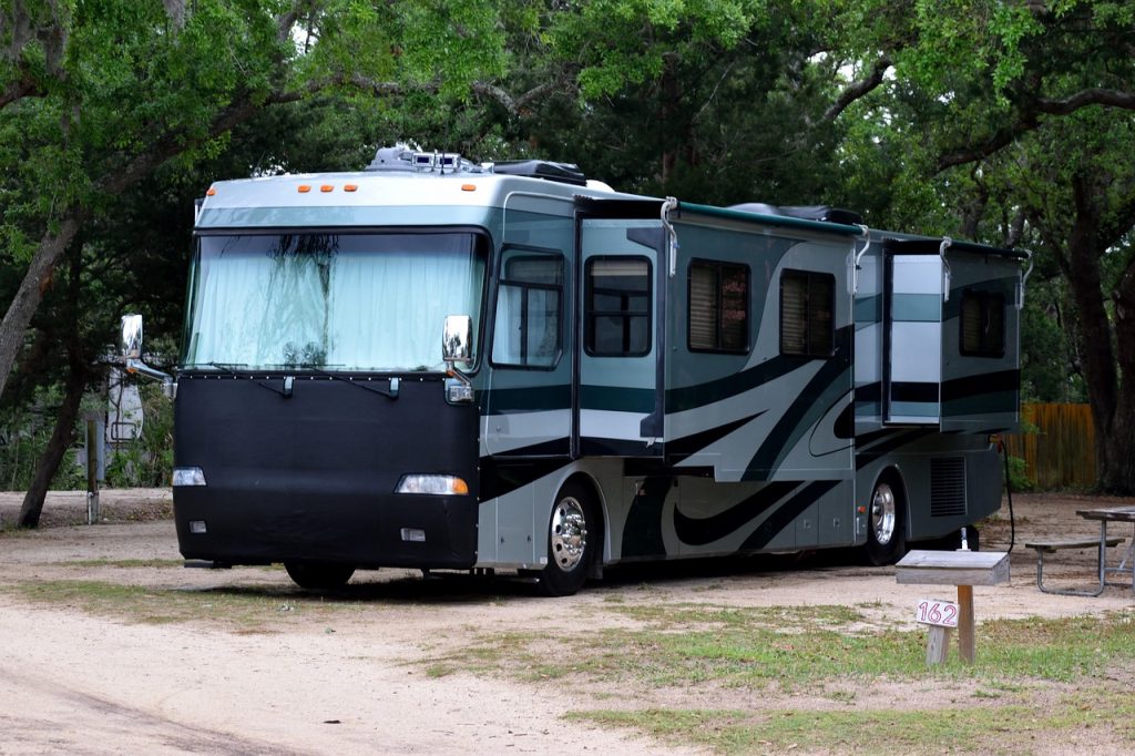 The image shows a modern Class A motorhome parked at a wooded campground with slide-outs extended, highlighting a common scenario where motorhome registration may still be required even if the vehicle is stationary.
