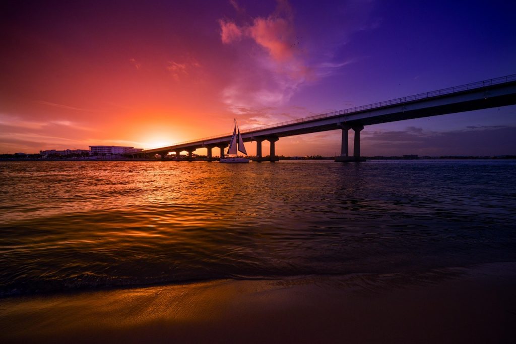 A sailboat glides beneath a bridge at sunset, where Alabama’s skies glow in shades of orange and violet over the water.