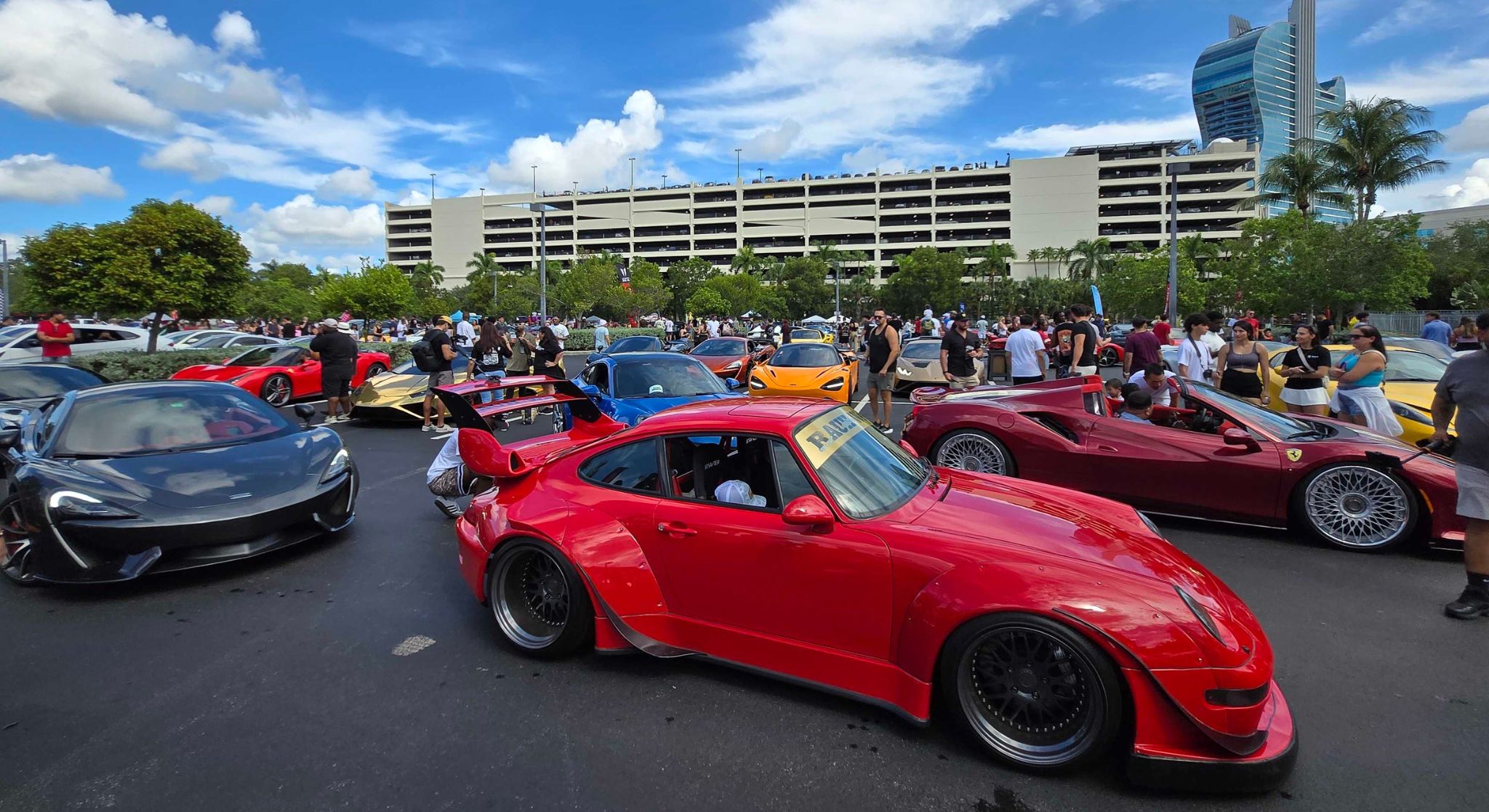 The image shows a line of luxury supercars parked side-by-side at an outdoor event, with palm trees and a clear sky in the background.