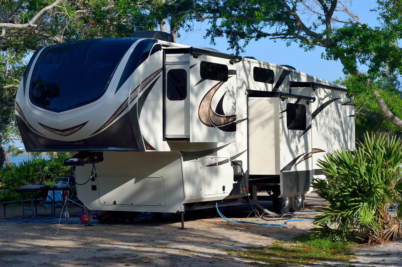 A large fifth-wheel RV with multiple slide-outs is parked at a shaded campsite, surrounded by trees and greenery.