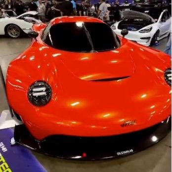 A sleek red race car with an aerodynamic design is displayed indoors at the SEMA auto show surrounded by other high-performance vehicles.