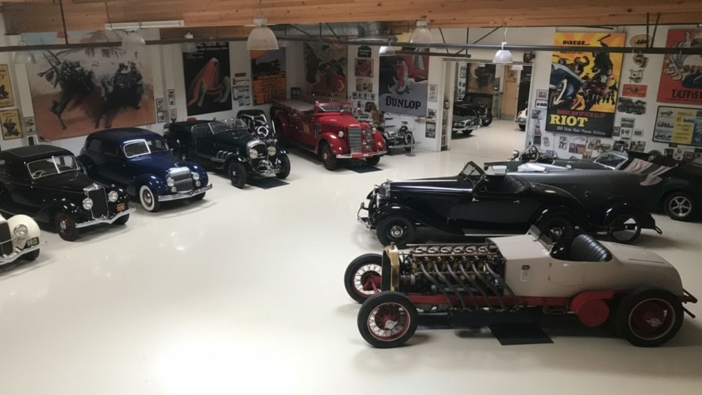 An expansive view inside Jay Leno's Garage, featuring a remarkable collection of pre-war and vintage automobiles, including classic luxury cars, a red antique fire truck, a unique multi-cylinder open-wheel race car, and various roadsters, amidst a backdrop of retro automotive posters.