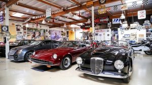 Jay Leno's Garage interior showcasing a diverse collection of classic cars and modern vehicles, including a Cadillac CTS-V, a red classic coupe, and a black vintage convertible, parked in a workshop adorned with automotive memorabilia.