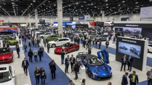 High-angle view of a large international auto show floor with luxury sedans, SUVs, and sports cars on display with a crowd of attendees.