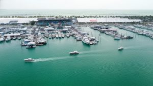 An aerial perspective of the Miami International Boat Show featuring hundreds of boats and yachts docked at a massive temporary pier structure with white event tents along the shoreline and boats cruising in the green water.