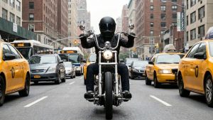 Front-facing view of a rider in a black helmet and leather jacket on a chrome custom chopper, cutting through heavy New York City traffic flanked by yellow taxis and city buses