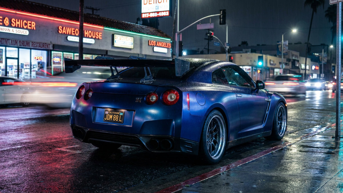 Rear view of a matte blue Nissan GT-R with a large rear wing and California license plate, sitting at a rain-soaked Los Angeles intersection at night with neon storefronts and streaking traffic lights in the background